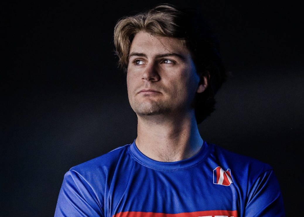 A man with light brown hair wearing a blue athletic shirt stands against a dark background, looking to the side with a neutral expression, reminiscent of Colton Herta from the IndyCar series.