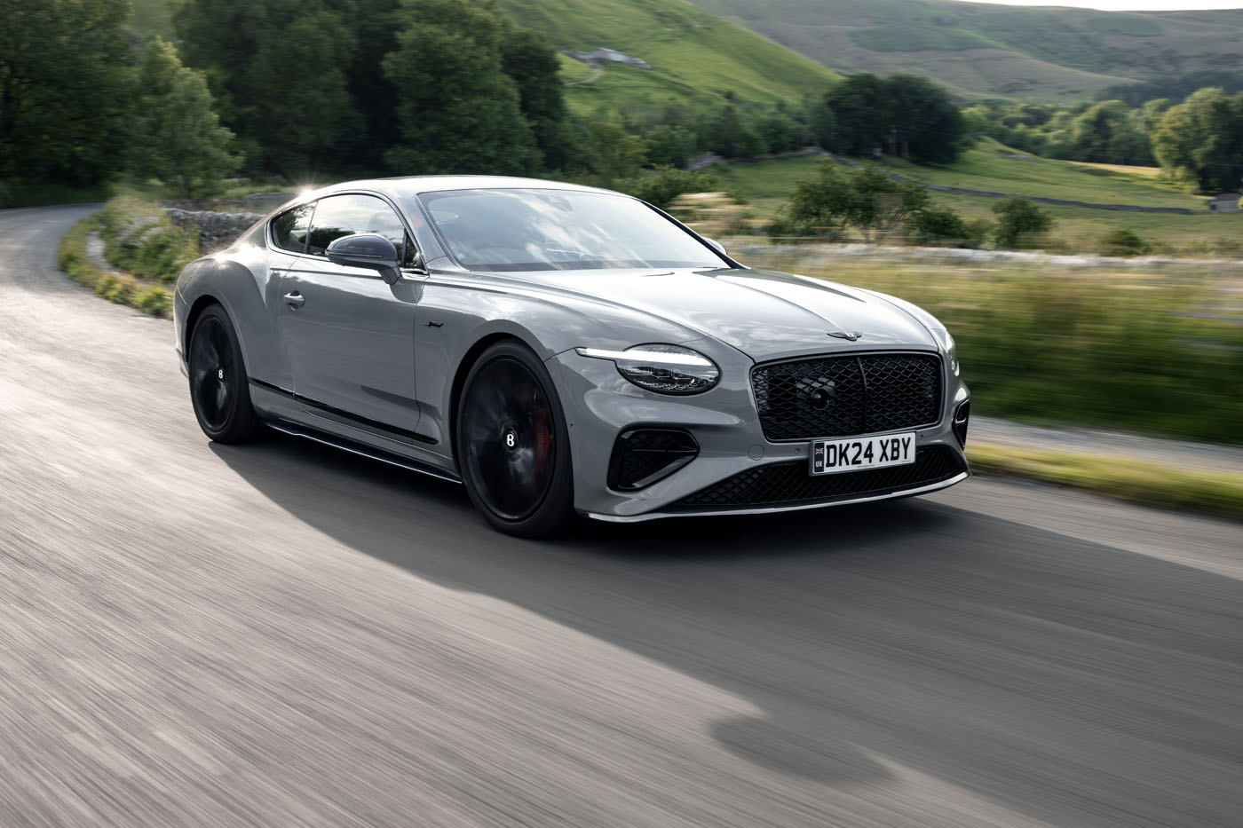 A silver Bentley coupe with a carbon fiber styling package glides along a winding rural road, surrounded by lush greenery and rolling hills in the daylight.