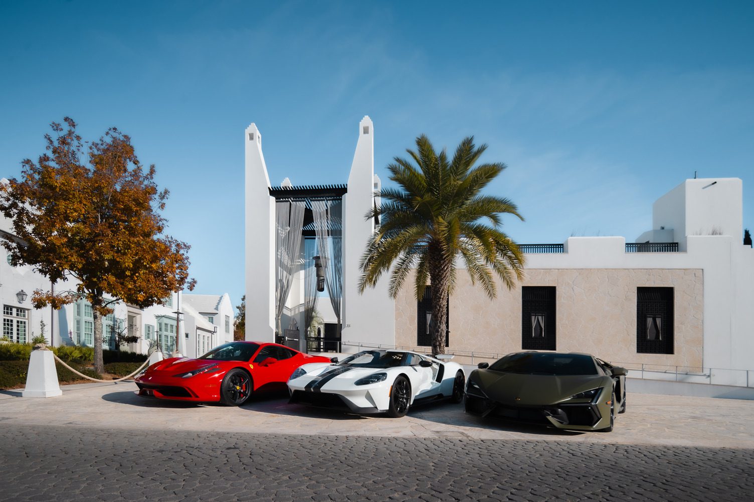 Three luxury sports cars—a red, a white, and a dark green—are parked side by side in front of a modern white building at Alys Beach, capturing The Art of the Drive beneath swaying palms and a clear blue sky.