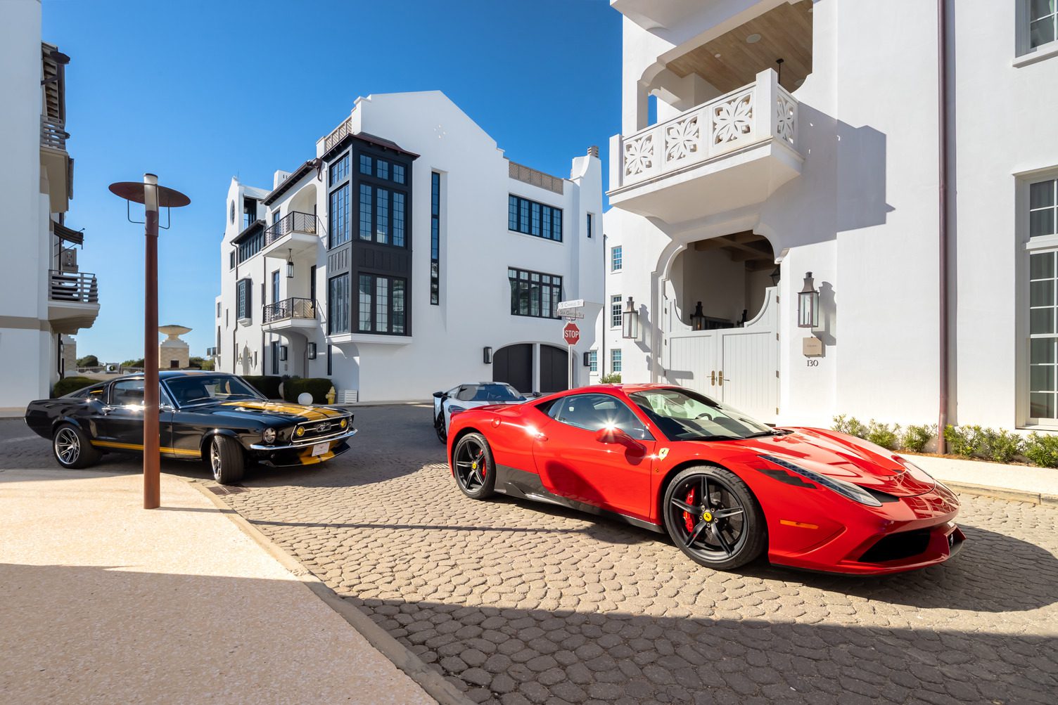 A red Ferrari and a black classic car parked on a cobblestone street in Alys Beach showcase the Art of the Drive, set against modern white buildings beneath a clear blue sky—signature moments from Cars of 30A.