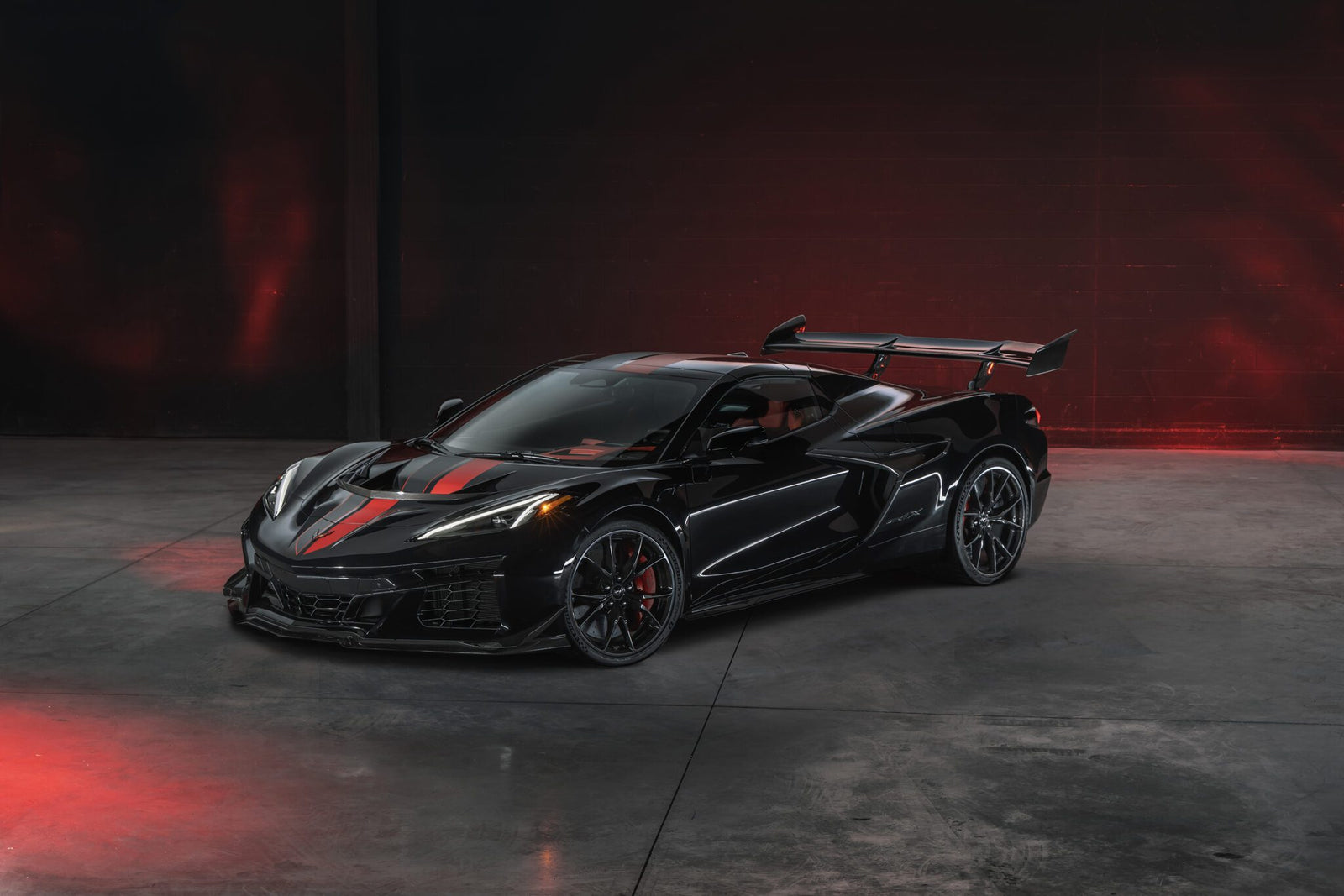 A black Chevrolet Corvette ZR1X hypercar with red accents and a large rear wing is parked in an empty, dimly lit garage with striking red lighting effects.