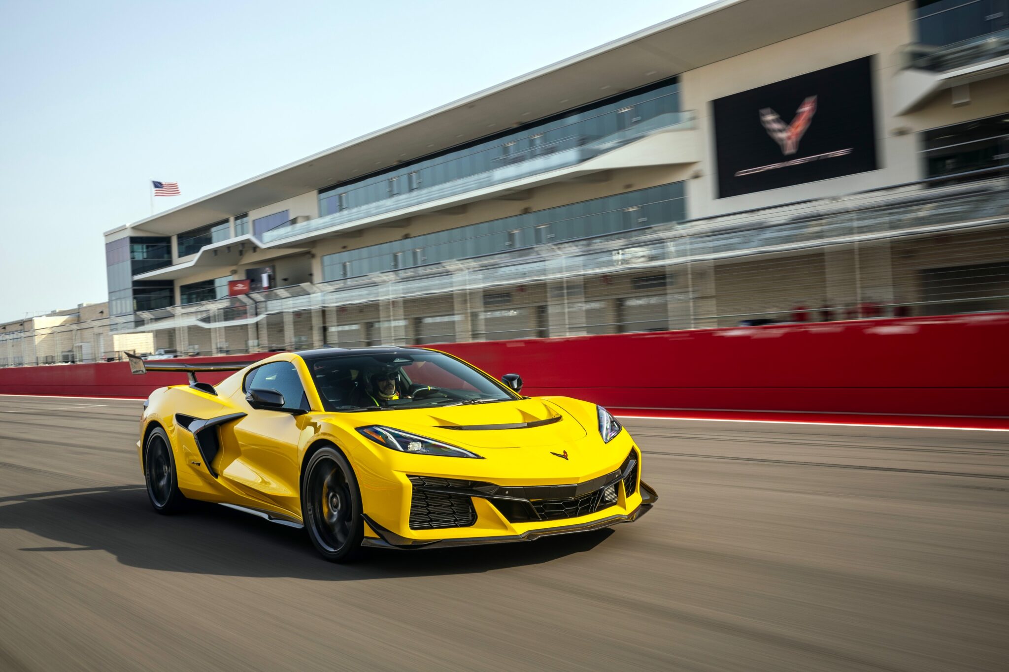 A yellow Track Review Corvette ZR1, proving 1000 HP doesn’t need a seven-figure price, speeds along a racetrack in front of a modern, multi-story building with large windows and a red barrier.