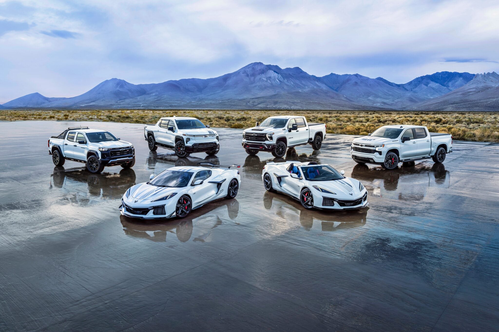 Six white vehicles, including three pickup trucks and three sports cars from the Chevy Honors America’s 250th Anniversary Ultra-Exclusive Stars & Steel Collection, are parked on a wet salt flat with mountains in the background under a cloudy sky.