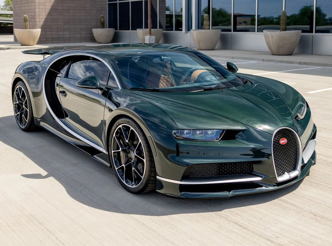 A dark green Bugatti Chiron hypercar is parked outdoors on a concrete surface near a modern building with large windows and potted plants.