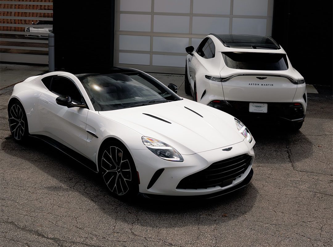 Two white Aston Martin cars, representing Cleveland Motorsports’ exquisite selection of exotic cars, are parked on cracked pavement in front of a closed garage door—one seen from the front, the other from the rear.