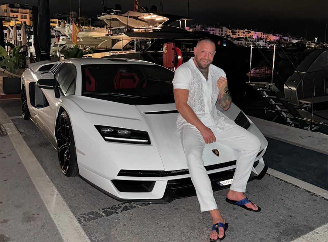 A man in white clothing sits on a white Lamborghini Countach, part of an impressive car collection, parked near a marina with boats in the background at night.