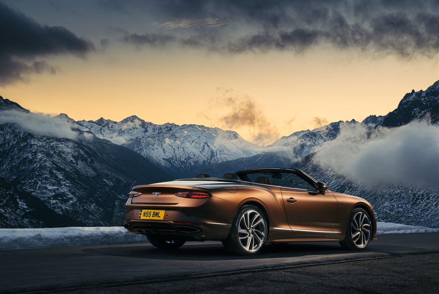 A brown Bentley Continental GT Speed is parked on a mountain road at sunset, overlooking snow-covered peaks and a cloudy sky, dreaming of the drive ahead powered by cutting-edge eFuels.