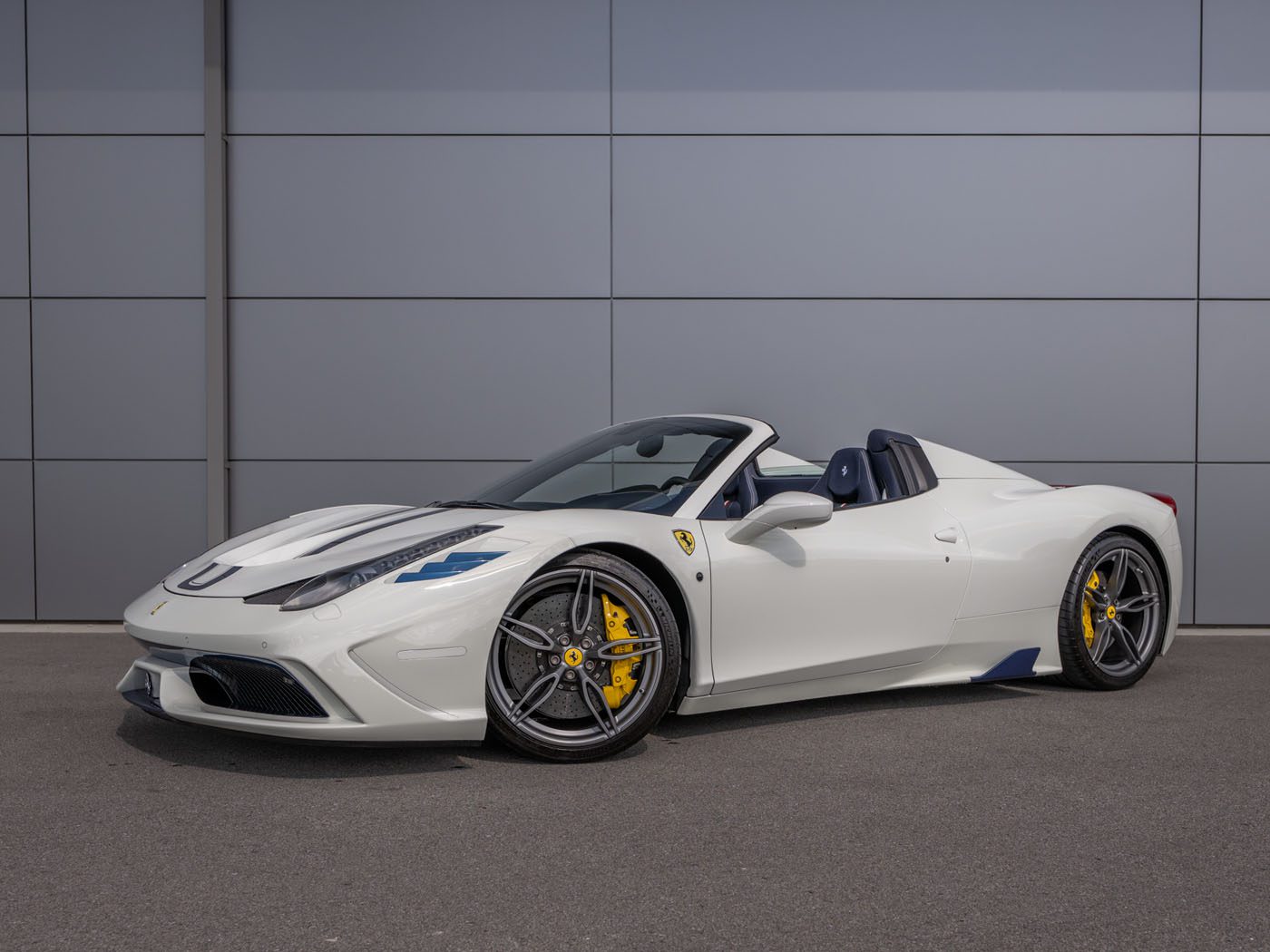 An ultra-rare Ferrari 458 Speciale Aperta, a white convertible sports car with yellow brake calipers, is parked on a paved surface in front of a gray paneled wall.