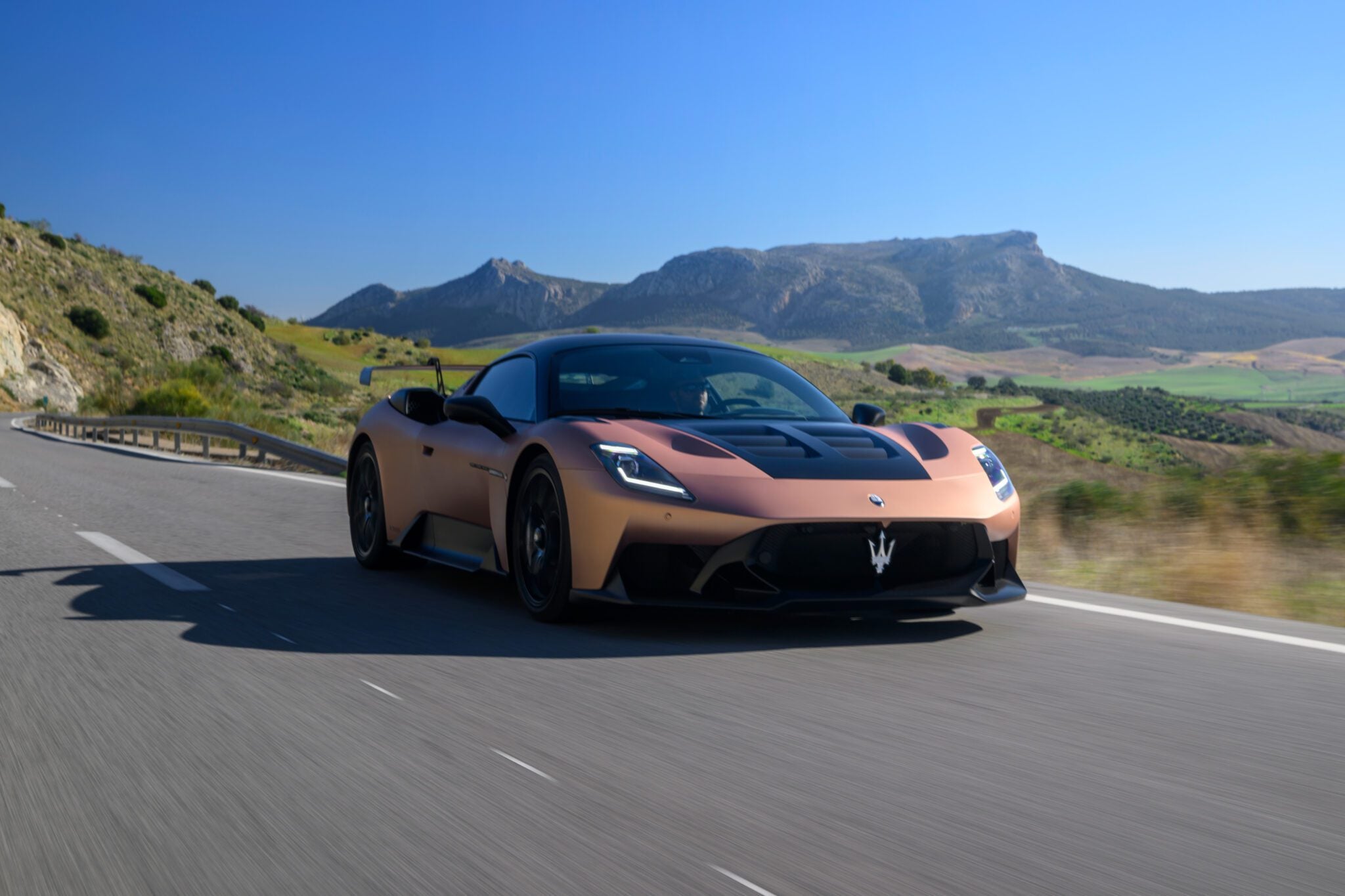 A sleek Maserati sports car races along a highway with mountainous scenery in the background under a clear blue sky.
