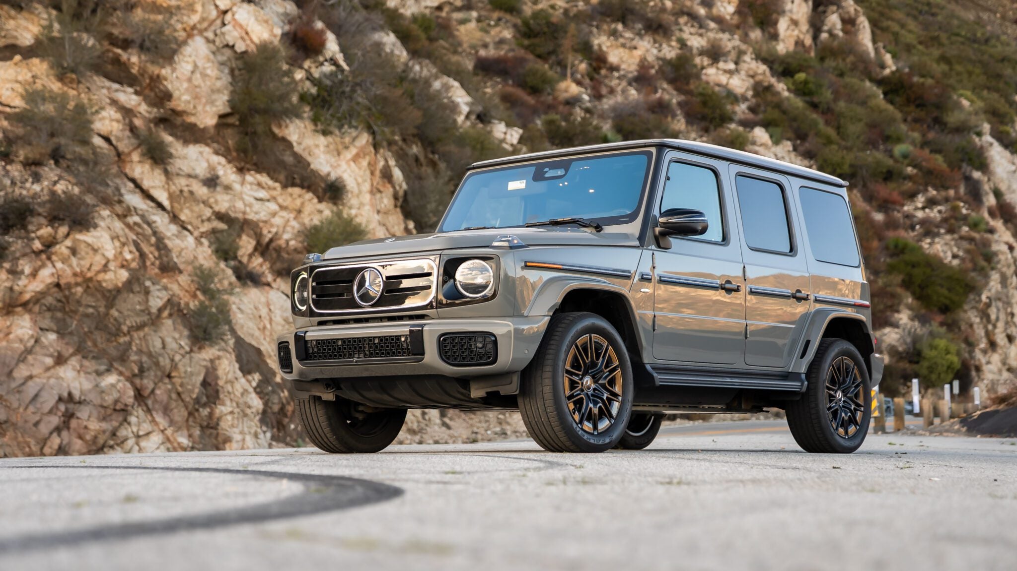 A silver Mercedes-Benz G-Class SUV, a true Mercedes icon, is parked on a paved road with rocky hills and greenery in the background.