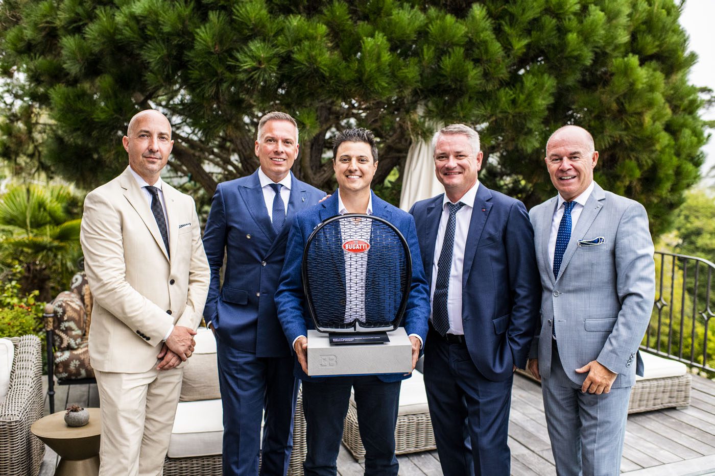 Five men in suits stand outdoors, posing for a photo. Abraham Cohen, at the center, holds a Bugatti Broward trophy for Best Performing Brand Manager. Green foliage and outdoor furniture are visible in the background.
