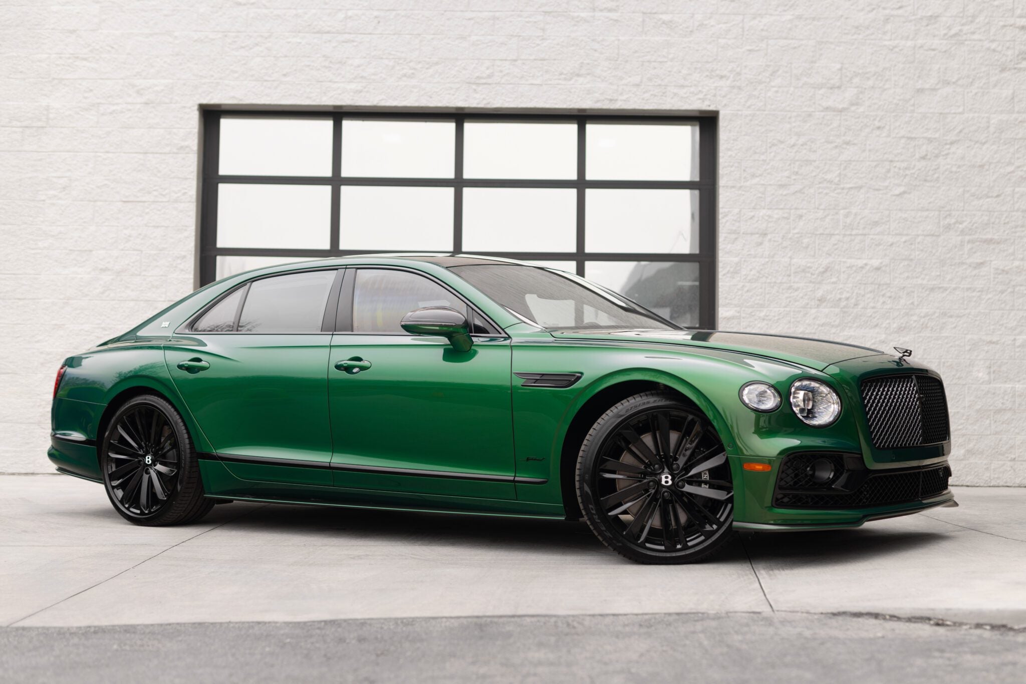 A green Bentley Continental GT End of Era Edition luxury sedan parked on a concrete driveway in front of a large window with a black frame.