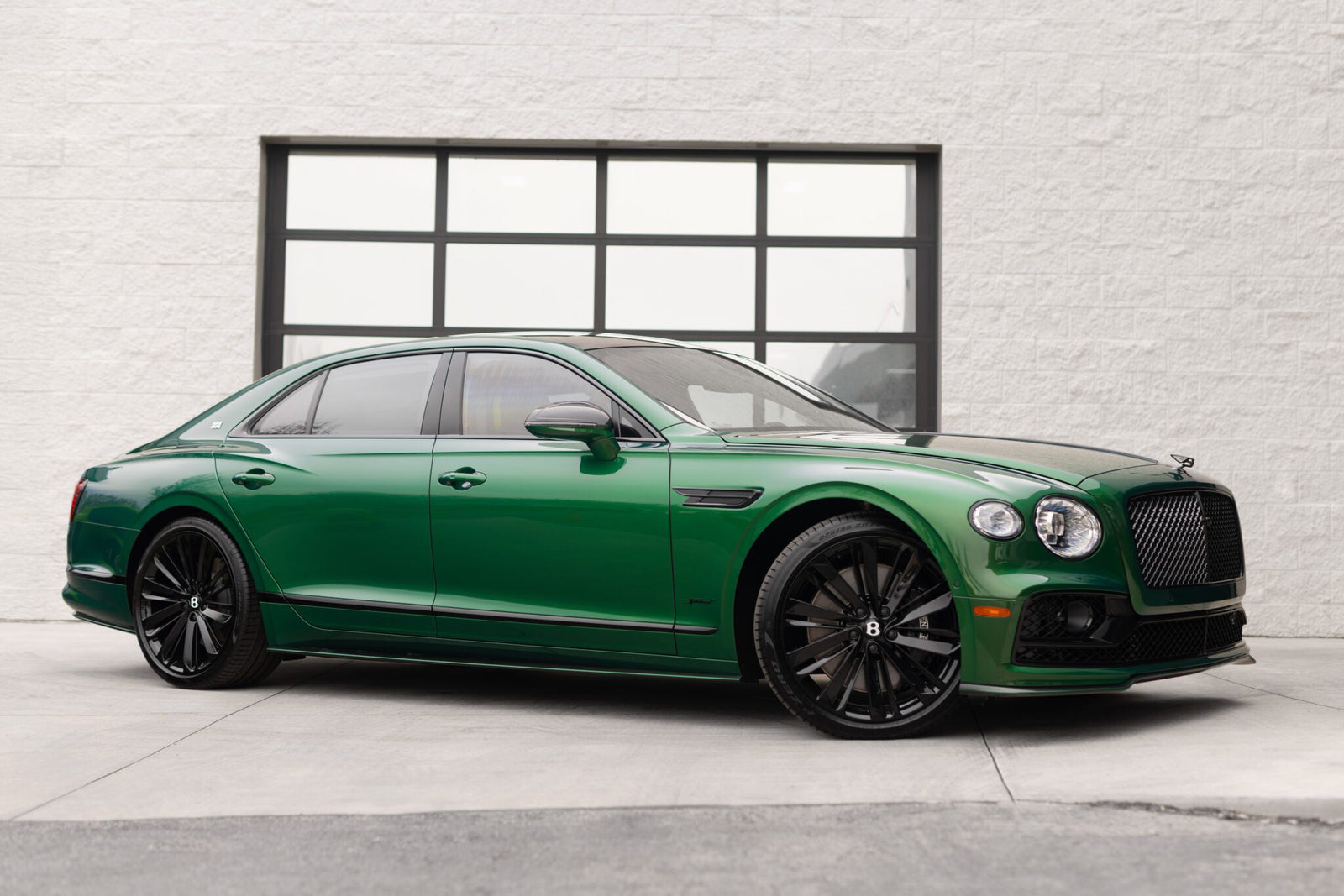 A green Bentley Continental GT End of Era Edition luxury sedan parked on a concrete driveway in front of a large window with a black frame.