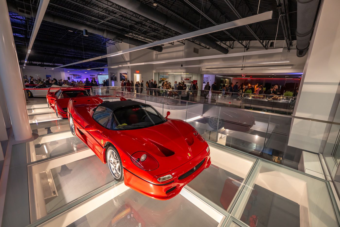 A red sports car is displayed on a glass floor in a modern Nashville showroom, with people from the Ferrari community gathered in the background for a Make-A-Wish event.