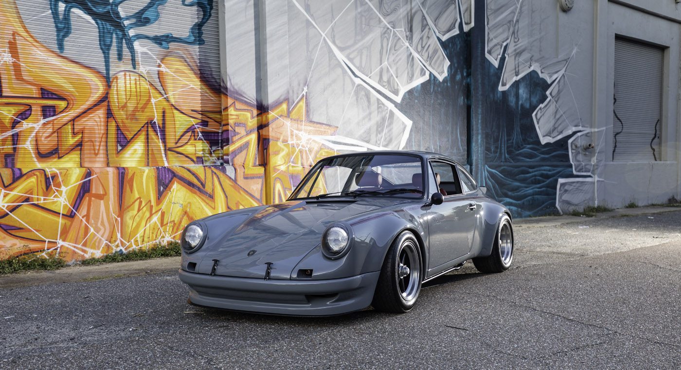 A gray classic sports car from Classic 9 Motorwerks is parked on a street in front of a building with colorful graffiti and a monochrome mural, evoking the vibrant energy of an exotic car festival.