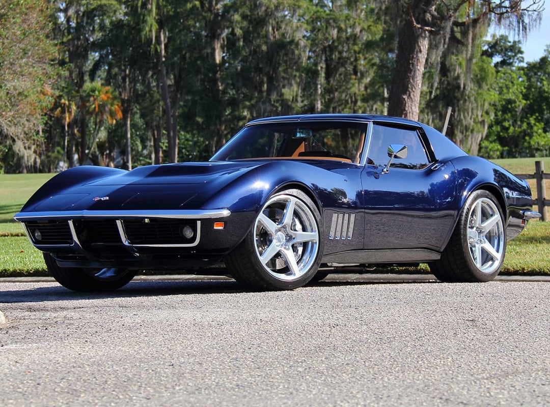 A dark blue Corvette Restomod classic Chevrolet sports car is parked on a paved road with trees and grass in the background, as seen at Mecum Kissimmee supporting Curing Kids Cancer.