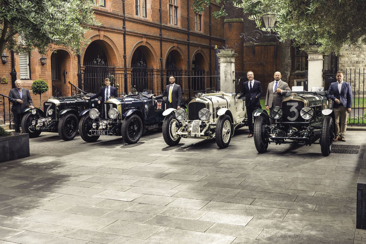 Six men in suits stand beside four vintage cars parked in a courtyard with red brick buildings and stone pavement in the background.