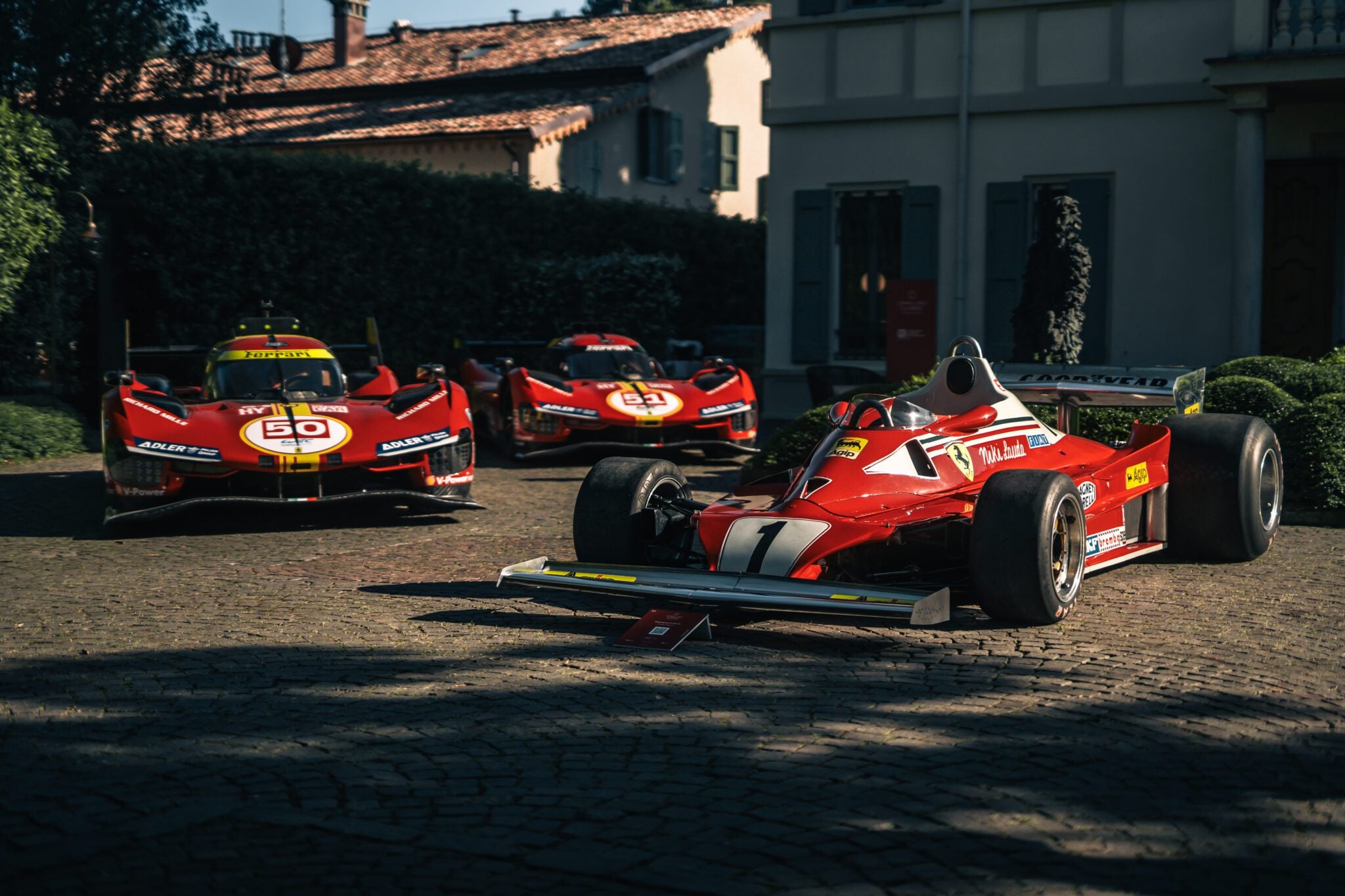 Three red Ferrari race cars, including Best of Show Winners from the Cavallino Classic Modena 2025, are parked on a stone driveway in front of a house in daylight.