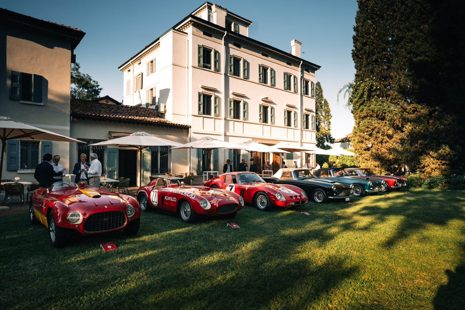 A row of classic sports cars is displayed on a lawn in front of a large, elegant white building at the renowned Cavallino Classic, with a few people standing nearby.