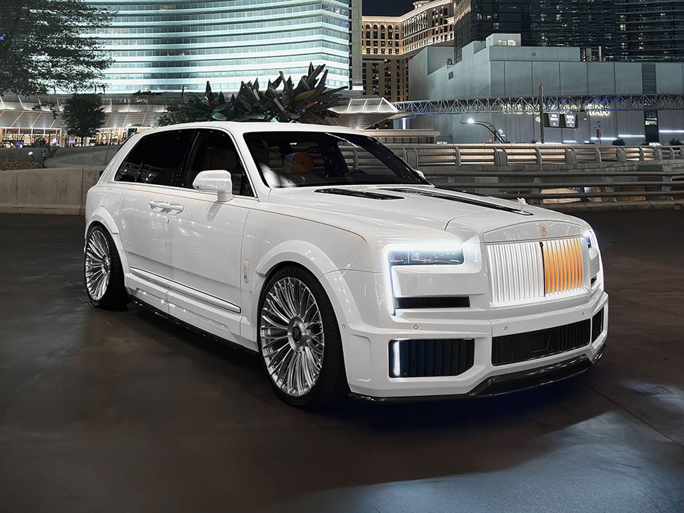 A white luxury SUV with custom wheels and an illuminated grille is parked on a city street at night, showcasing Urban Automotive style against the backdrop of modern buildings.