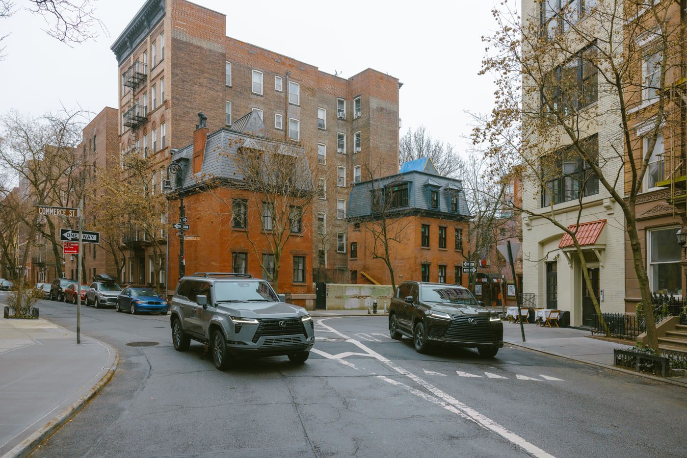 Two SUVs drive on a city street lined with brick apartment buildings, bare trees, and some parked cars on a cloudy day.