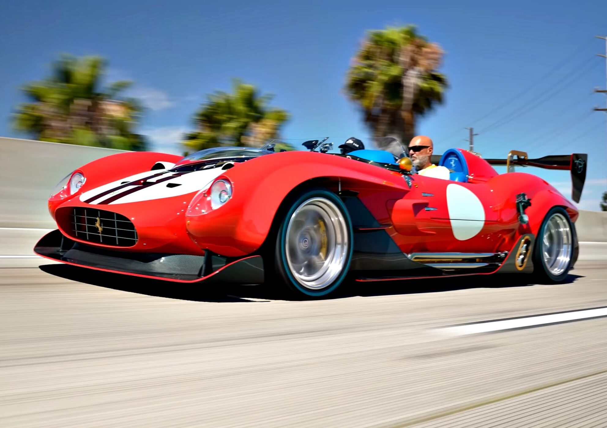 A red vintage-style Ferrari 612TR sports car with a white circle and black stripes is driven on a highway by a man wearing sunglasses; palm trees and blue sky in the background, capturing true V12 exotic flair.