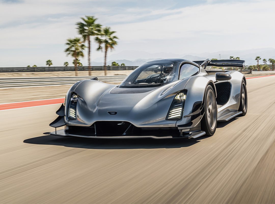 A silver American supercar drives at high speed on a racetrack with palm trees and mountains in the background.