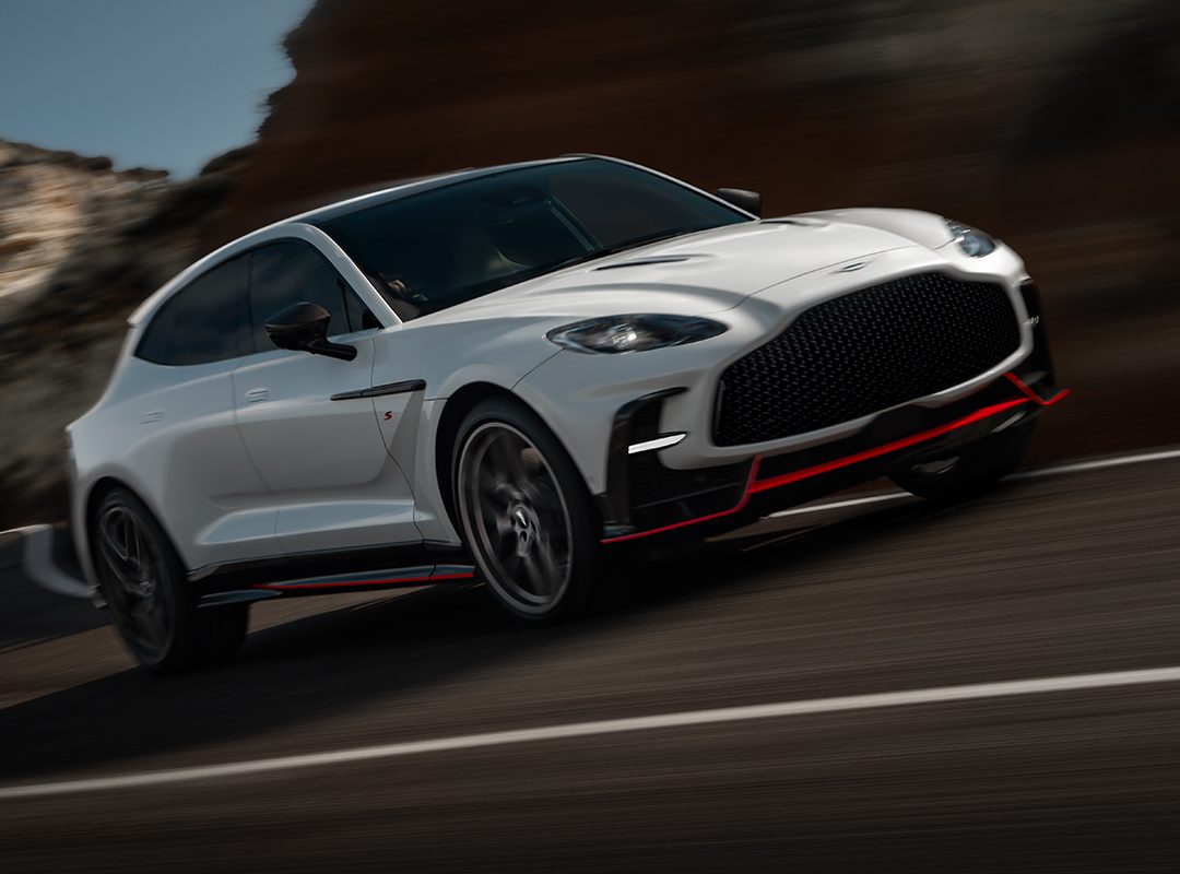A white Aston Martin DBX S Super-SUV with black and red accents is driving at speed on a curved mountain road.