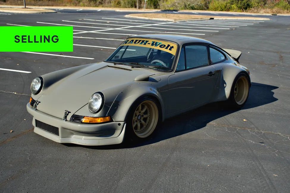 A matte grey modified Porsche 911 RSR with wide fenders and gold wheels is parked in an empty lot; a green "SELLING" banner appears in the top left corner, evoking the excitement of car auctions.