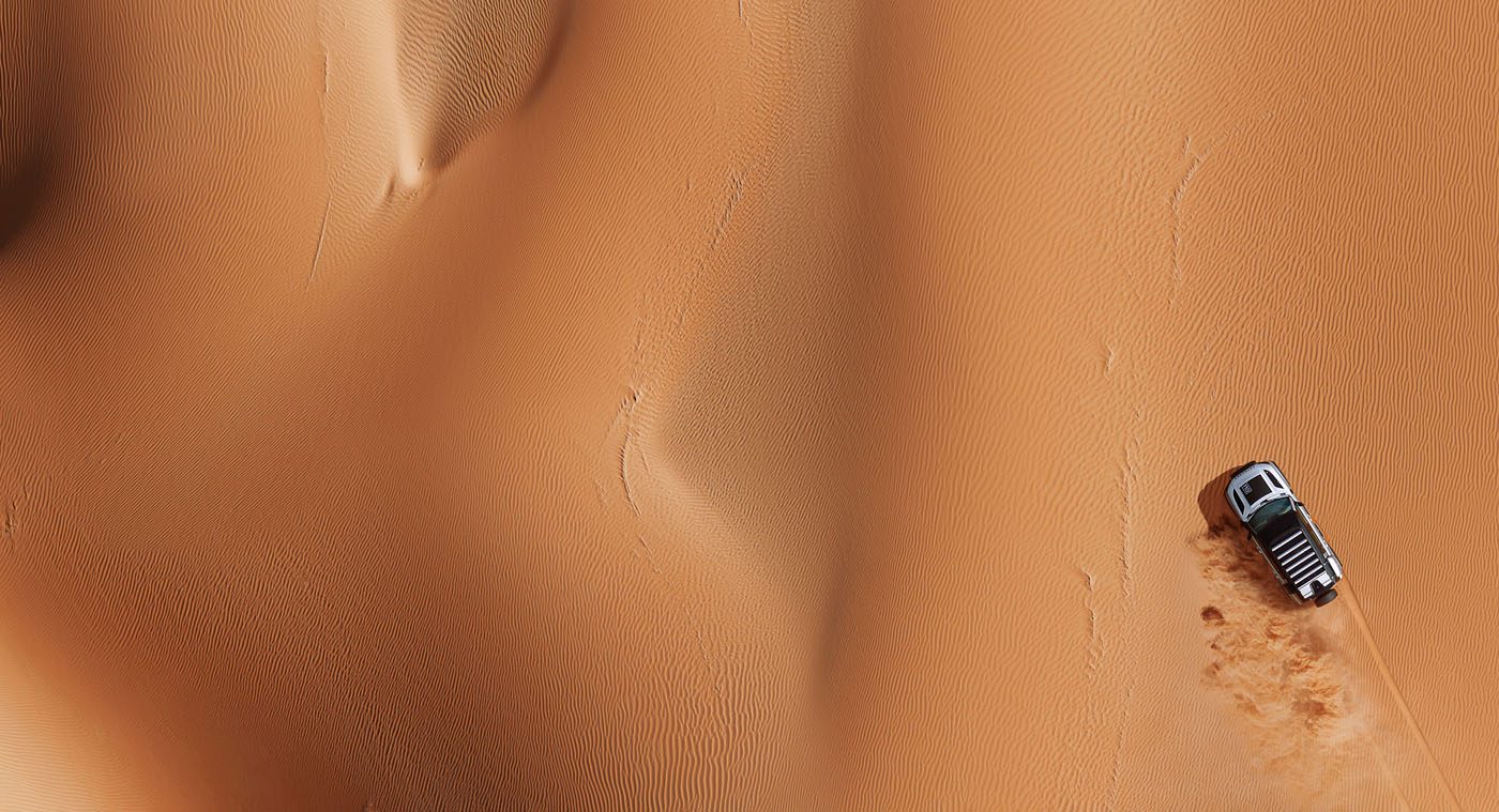 Aerial view of a Defender racing across vast orange sand dunes in a desert landscape, reminiscent of the thrilling Dakar Rally.