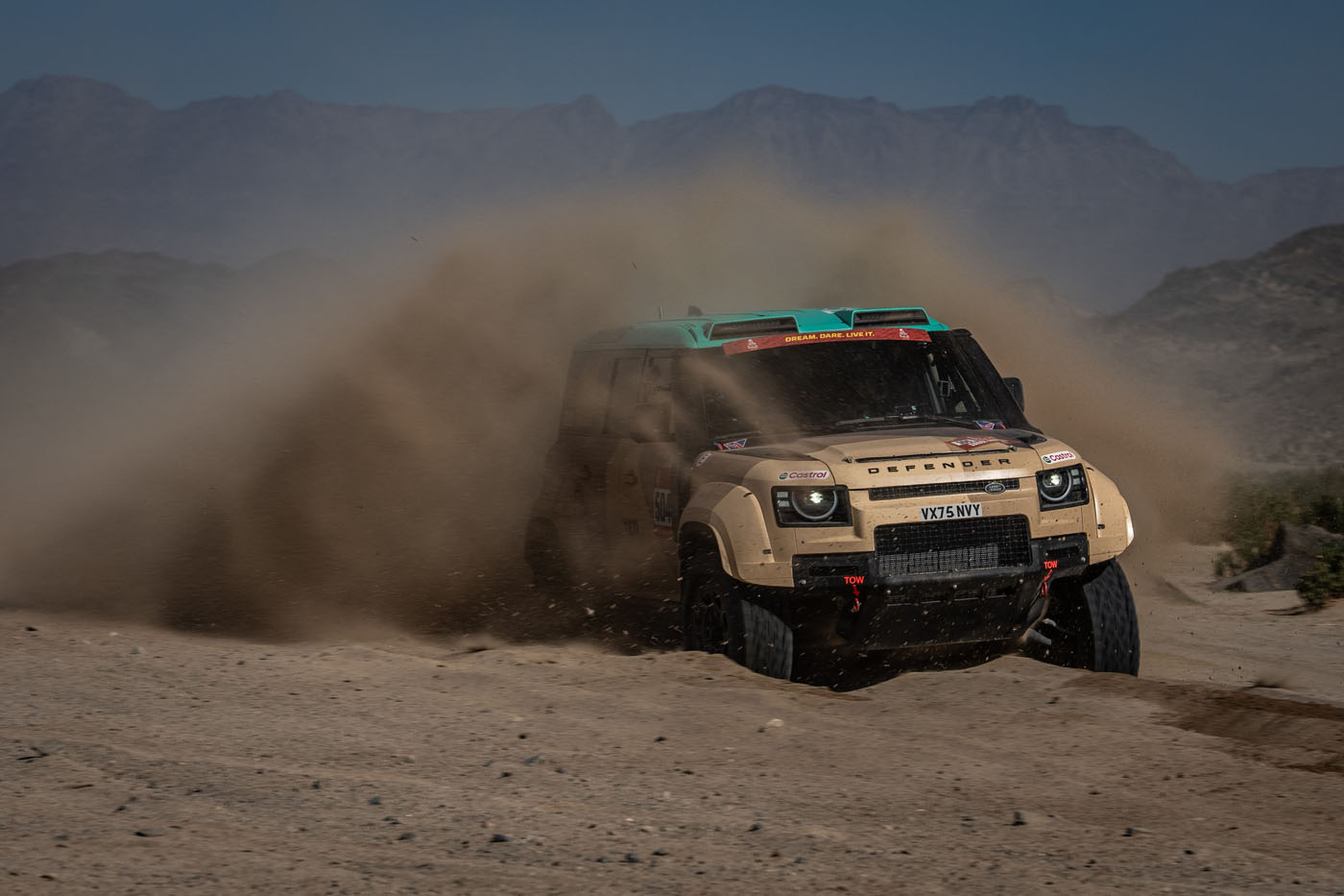 A beige D7X-R off-road vehicle speeds through a dusty desert trail, kicking up a large cloud of sand, with mountains visible in the background.