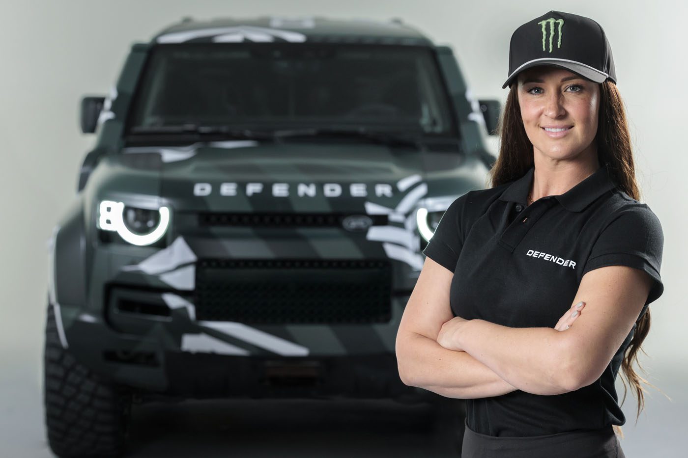 Sara Price stands confidently with arms crossed in front of a green Land Rover Defender, ready to take on any challenge like the Dakar Rally.