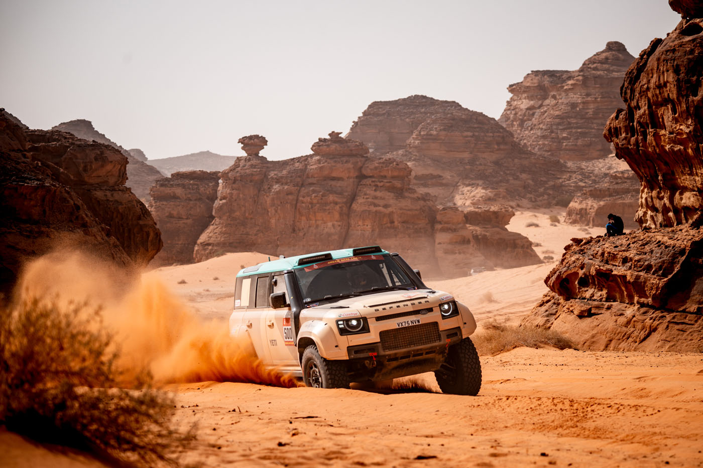 A white Defender Rally vehicle speeds through a sandy desert, kicking up orange dust, with rocky formations and a distant figure in the background—capturing the thrill of Stage 3 of the Dakar Rally.