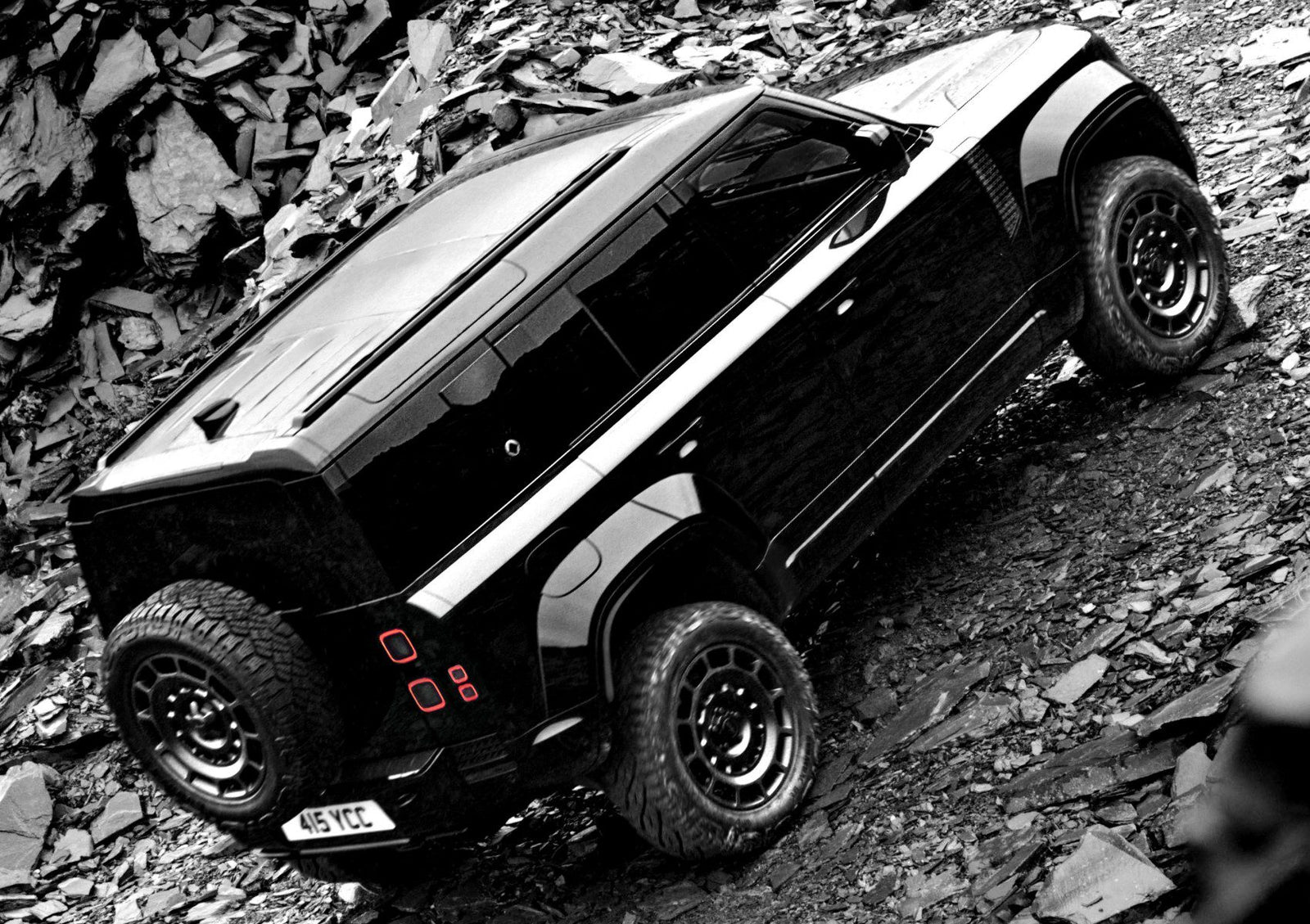 A 2026 Land Rover Defender in OCTA Black with a spare tire mounted on the rear drives up a rocky, uneven slope.