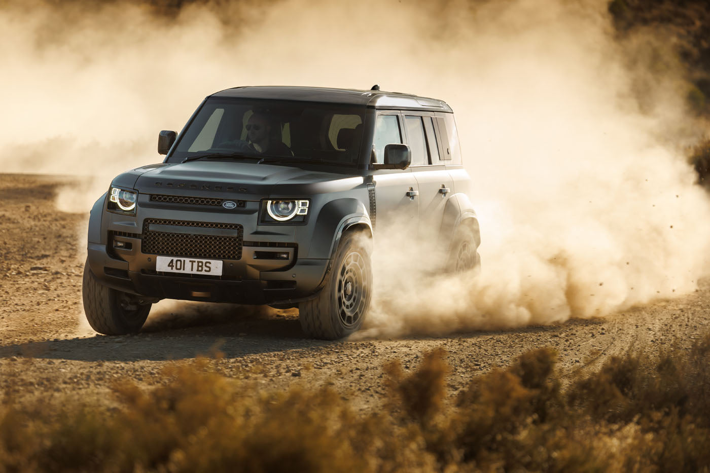 A dark gray Defender SUV drives through a dusty, rugged terrain, stirring up clouds of dust. Resembling the powerful Dakar Rally cars, the vehicle's headlights are on as it moves toward the left.
