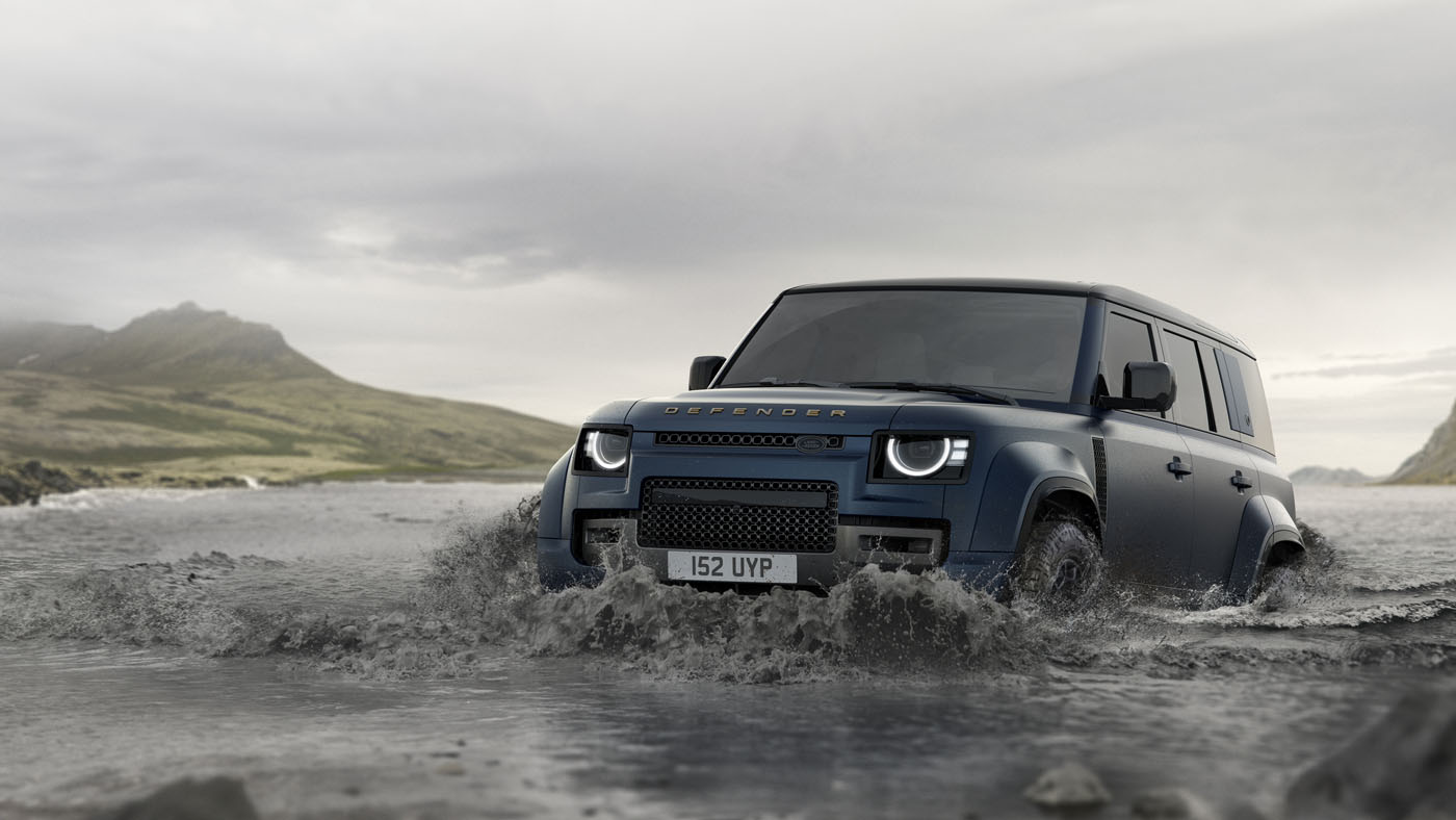 A blue Land Rover Defender showcasing its latest design updates drives through shallow water, set against a backdrop of mountains and cloudy skies.