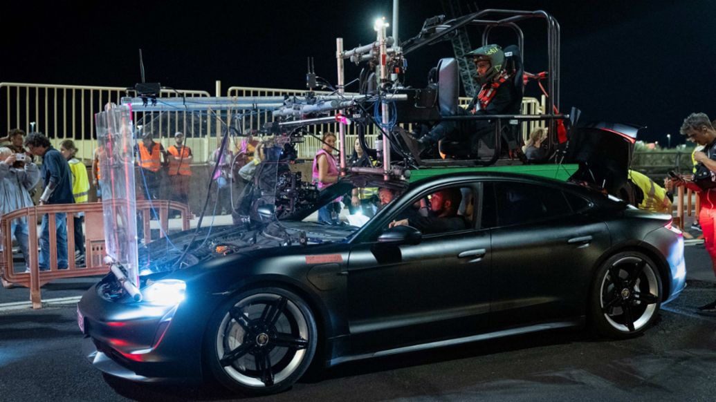 A camera rig is securely mounted on a sleek Porsche Taycan, with a crew member seated atop during an intense nighttime film shoot for Den Of Thieves 2, surrounded by bustling crew members in the background.