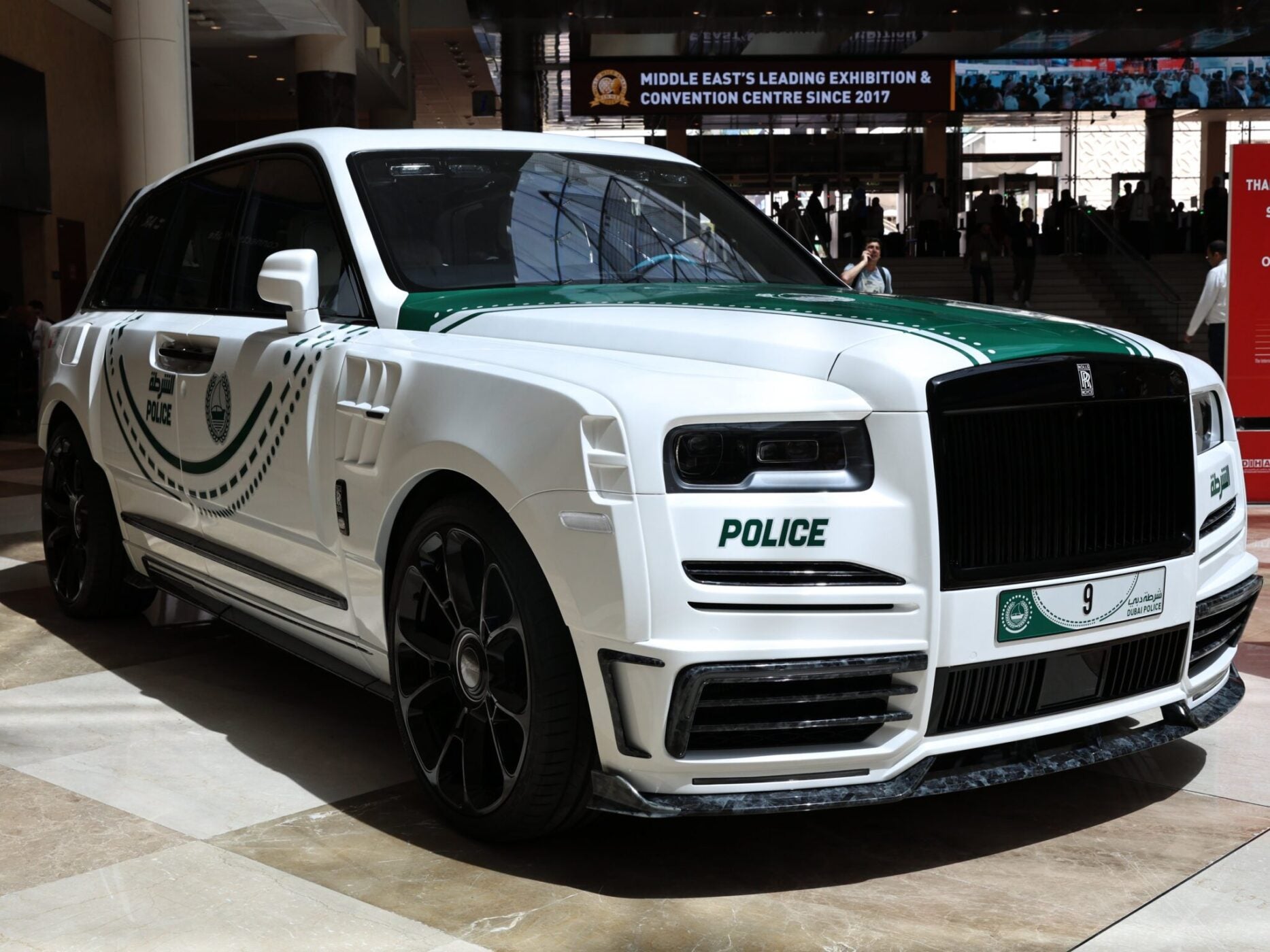A white Rolls-Royce SUV with green police markings and emblems is displayed indoors, featuring a Dubai police license plate.