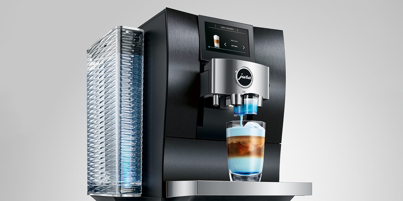 The JURA Z10 in a new color dispenses a beautifully layered latte into a clear glass, showcasing its modern automatic features, sweet enhancement technology, digital display, and transparent water reservoir.