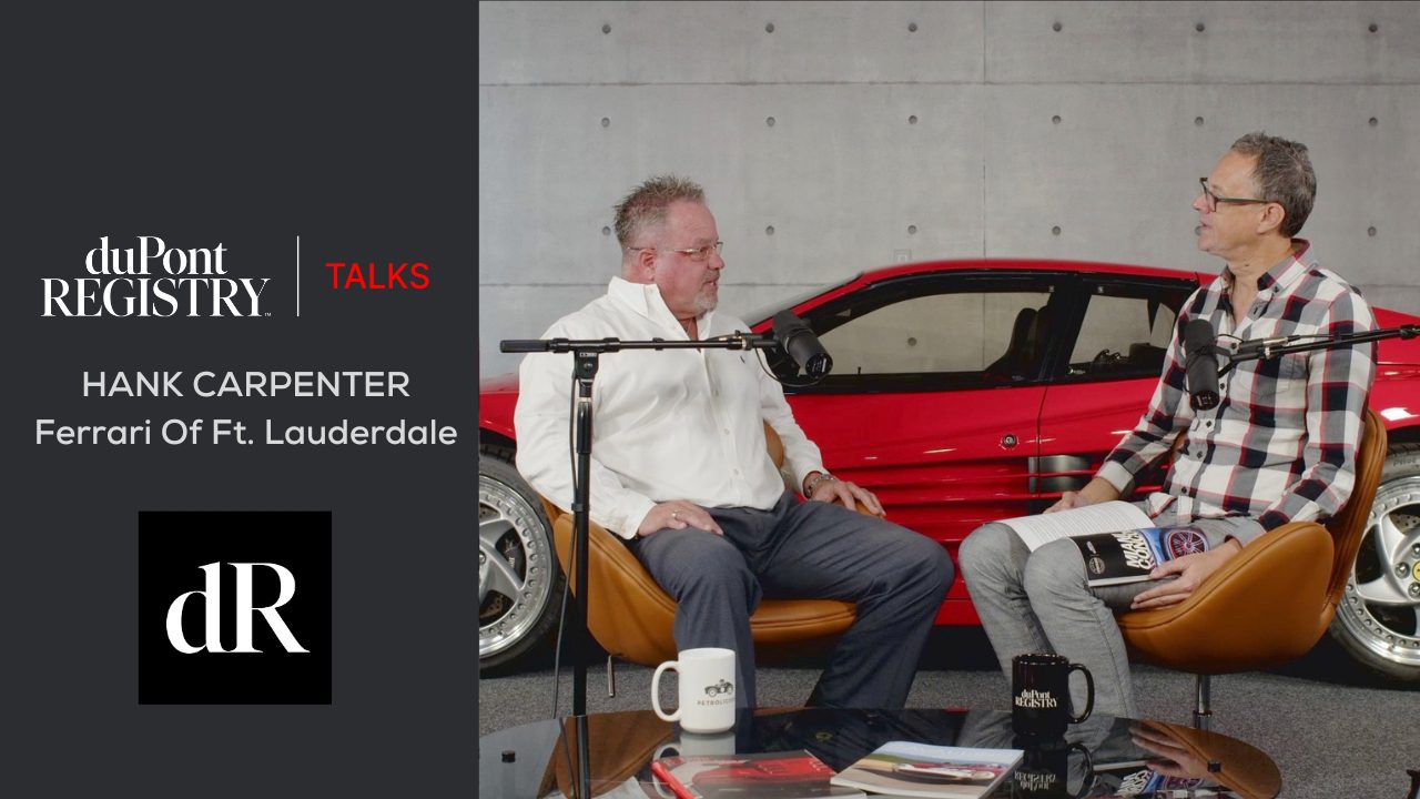 Two men seated and talking on a podcast set with microphones, with a striking red Ferrari in the background. Coffee mugs, magazines, and a duPont REGISTRY issue lie on the table, adding to the ambiance of this engaging discussion.