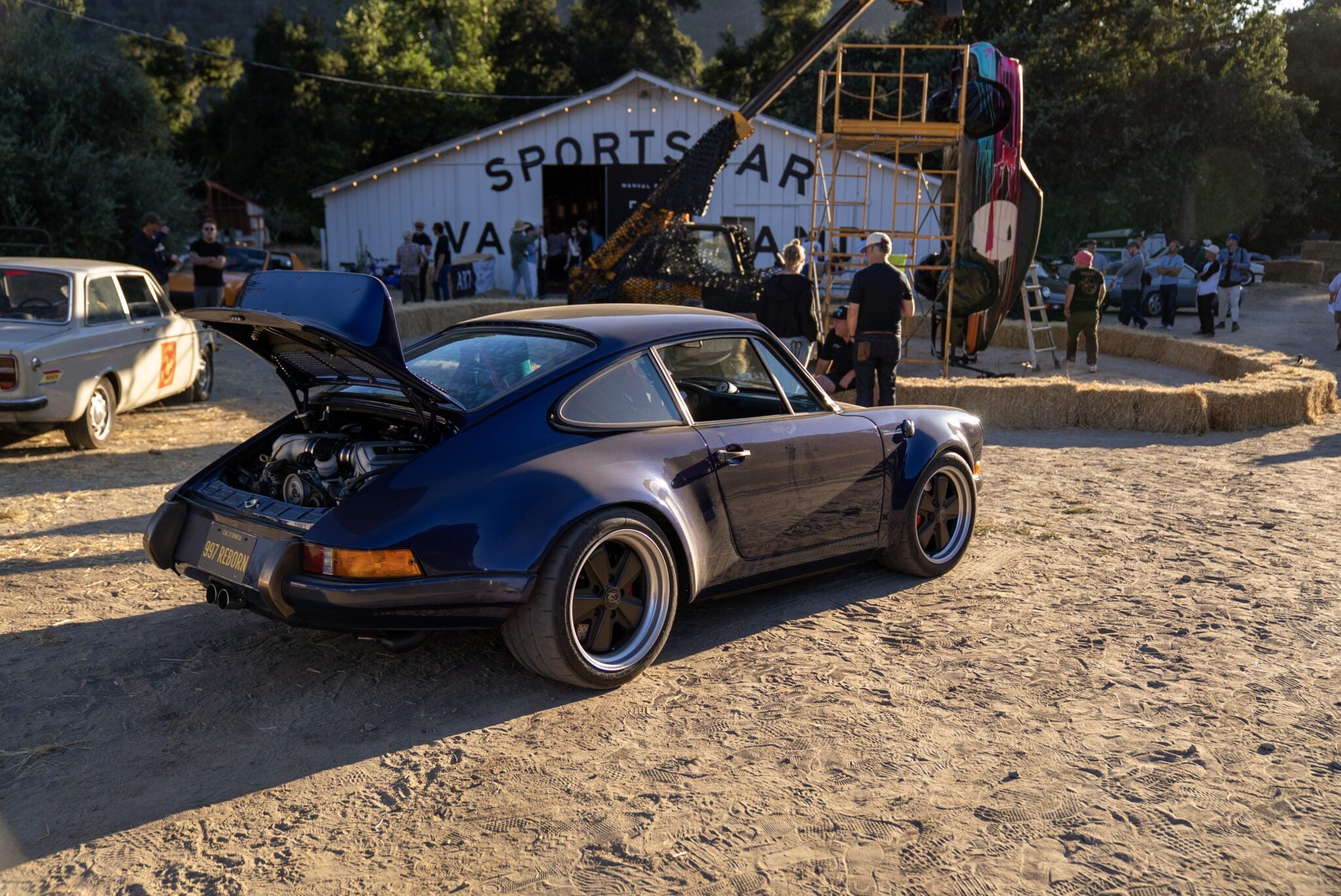 A blue classic Porsche with its rear trunk open is parked on a dirt lot near a white building labeled "SPORTS CAR"—an ideal Off the Beaten Path stop during your Car Week Escape, with people and other vehicles in the background.