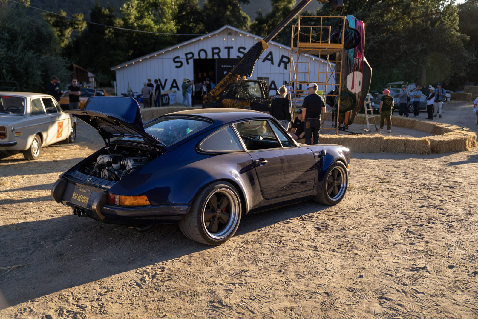 A blue classic Porsche with its rear trunk open is parked on a dirt lot near a white building labeled "SPORTS CAR"—an ideal Off the Beaten Path stop during your Car Week Escape, with people and other vehicles in the background.