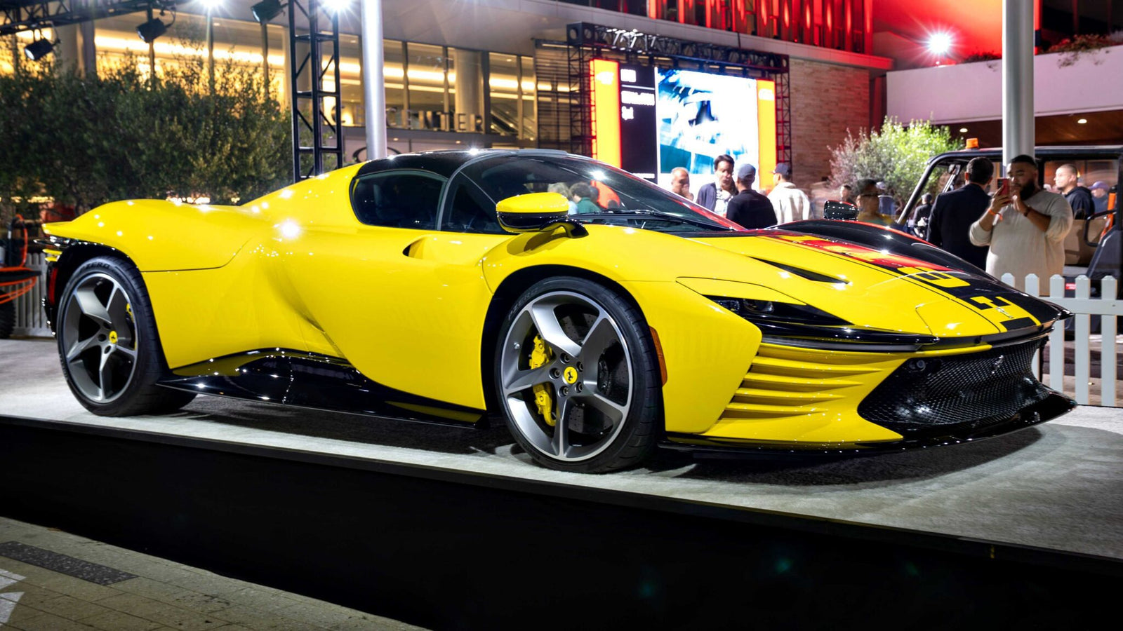 A bright yellow sports car, possibly one of the 10 Most Expensive Cars To Sell At 2025 Monterey Car Week Auctions, is displayed on a platform outdoors at night, with people and a building visible in the background.