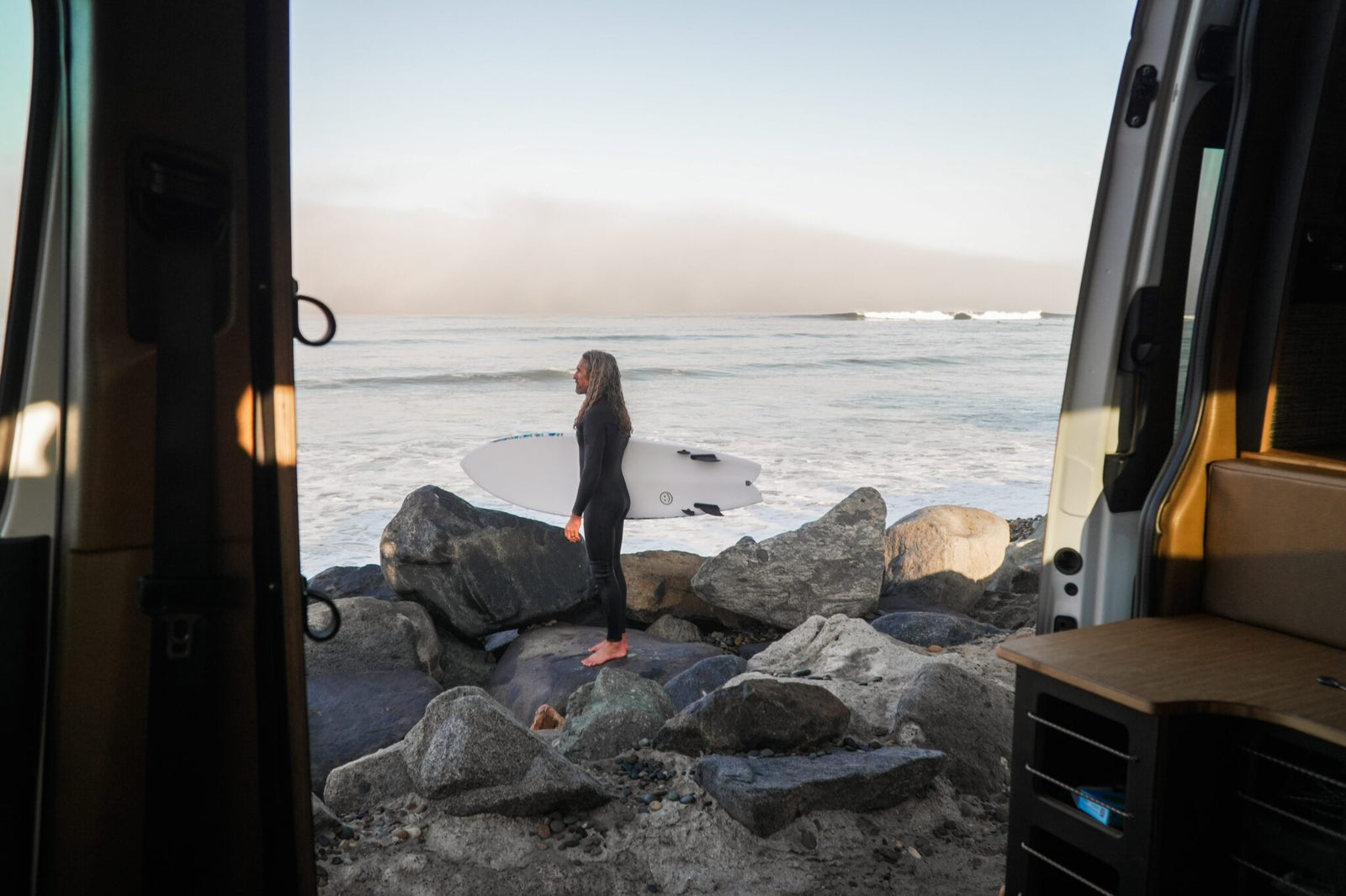 A person in a wetsuit stands barefoot on rocks by the ocean, holding a surfboard, viewed from inside a Rogue Van Company van.