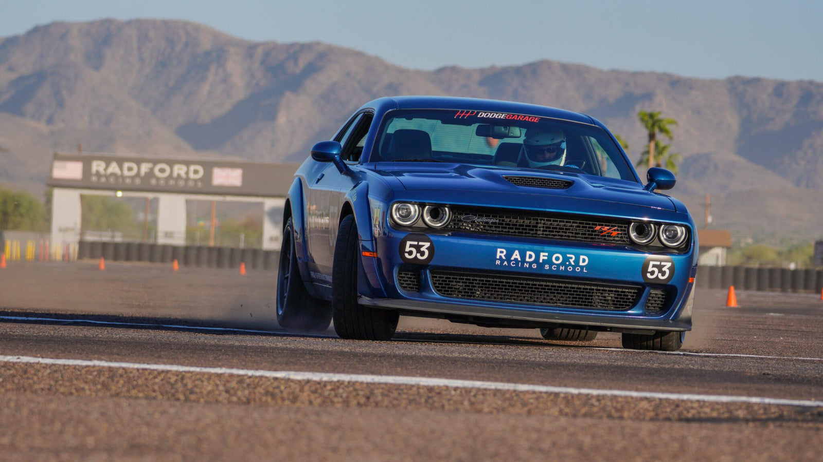 A blue Dodge sports car, number 53, races on a track at Radford Racing School, with mountains and the school's entrance sign visible in the background.