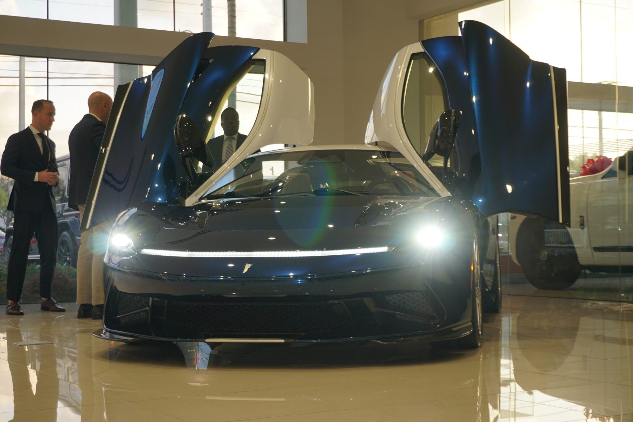 A luxury sports car with its doors open is displayed in Braman Motorcars' Palm Beach showroom. Nearby, three people stand conversing, perhaps captivated by the sleek design of Automobili Pininfarina.