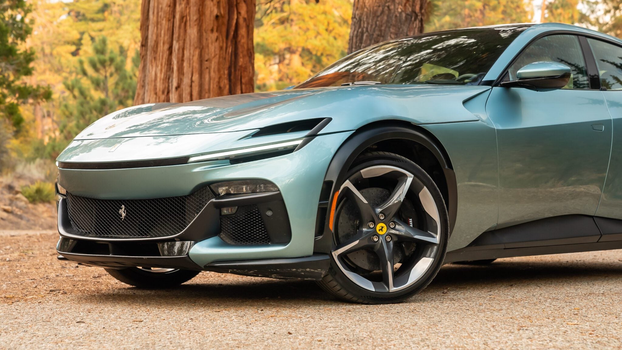 A sleek, light green V12 Grand Tourer sports car is parked on a dirt path, surrounded by tall trees. The iconic Prancing Horse emblem gleams proudly on its hood.