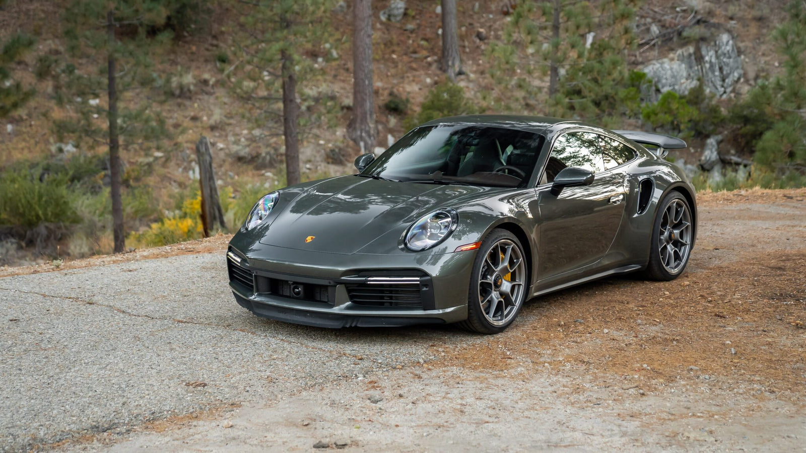 A gray sports car parked on a gravel path in a wooded area.