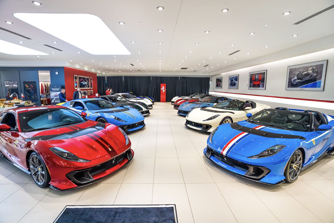A showroom featuring an exhibition of luxury sports cars, including the stunning Ferrari 812 Competizione in red, blue, and white. The well-lit room boasts framed photos on the walls, enhancing the elegant atmosphere.