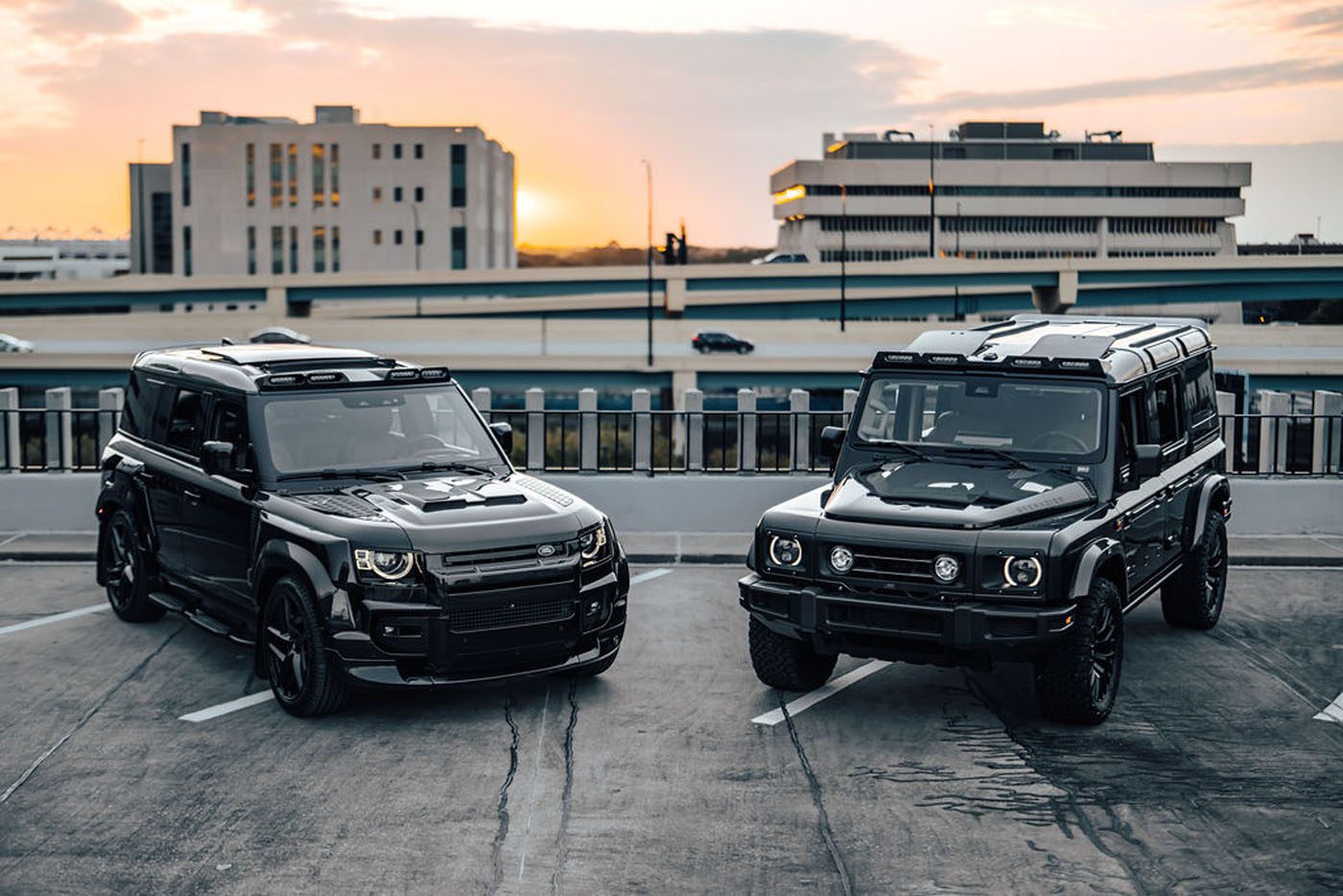 Two black SUVs—possibly Bespoke New Defenders—are parked on a rooftop parking lot at sunset, with multi-story buildings and a highway in the background.